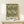 May include: Framed botanical poster with intricate green and cream acanthus leaf pattern in ornate Victorian style, displayed on light wood console table with small wrapped gift box beside it against neutral wall