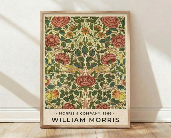 May include: Framed poster print featuring William Morris's ornate floral pattern from 1866, displayed in light wooden frame against white textured wall on wooden floor. Design shows symmetrical arrangement of deep red roses, green leaves, yellow birds, and orange flowers on cream background with "Morris & Company, 1866 - William Morris" text at bottom.