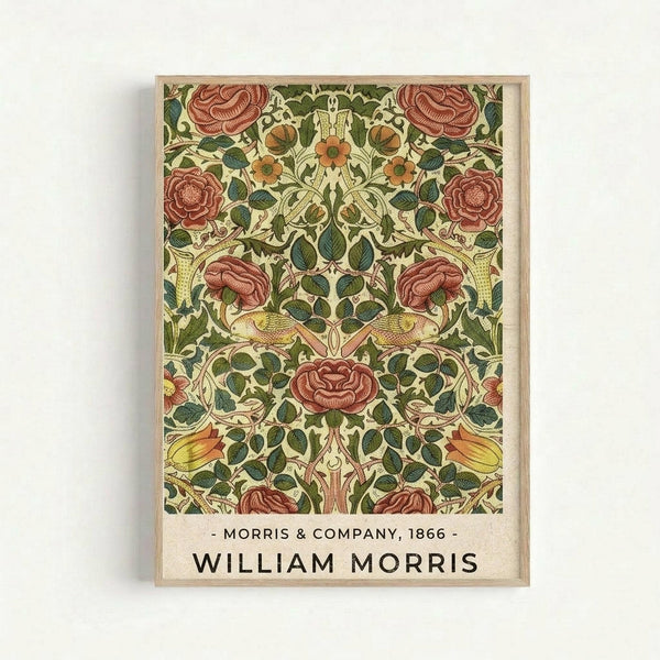 Framed vintage botanical poster featuring an ornate floral pattern with large red roses, orange flowers, and green foliage in William Morris's distinctive Arts and Crafts style. The symmetrical design shows intertwining vines and leaves on a cream background. Text at bottom reads "Morris & Company, 1866" and "William Morris" in black lettering. Natural wood frame hangs on white wall.