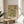 May include: Framed vintage botanical poster featuring William Morris design with coral roses, green leaves, and intricate floral patterns displayed on light wood round side table next to beige upholstered chair. Sunlight streams across neutral walls creating soft shadows. Plant leaves visible in corner of bright, minimalist room setting.