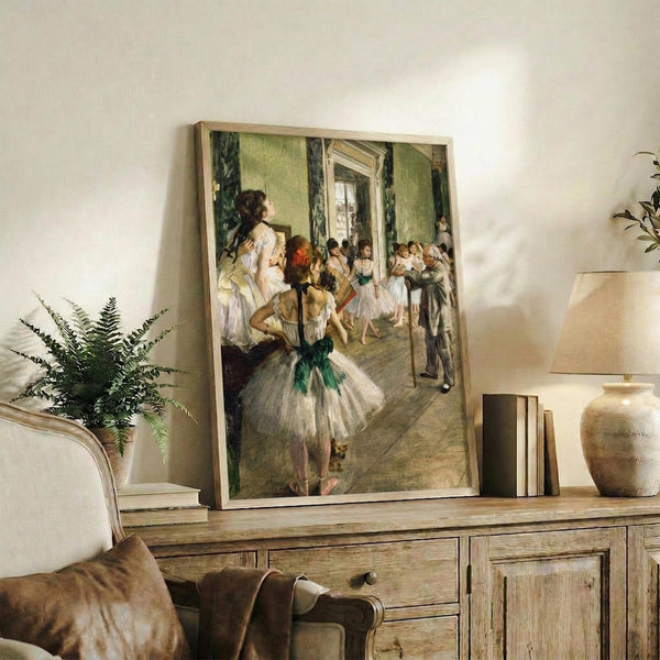 May include: Framed art print of ballet dancers in white tutus with green sashes practicing in an ornate studio with pale green walls and large mirror. Print displayed on rustic wooden sideboard alongside decorative pottery, books, fern plant, and cream upholstered chair with brown leather pillow visible in foreground.