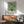 May include: Van Gogh roses poster print in wooden frame hanging above mid-century modern credenza with table lamp. Green fabric armchair with wooden arms sits nearby. Soft natural light creates shadows on white wall. Poster shows white roses in green vase against lime green background with artist name and date below artwork.