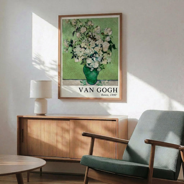 May include: Van Gogh roses poster print in wooden frame hanging above mid-century modern credenza with table lamp. Green fabric armchair with wooden arms sits nearby. Soft natural light creates shadows on white wall. Poster shows white roses in green vase against lime green background with artist name and date below artwork.