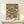 May include: Framed vintage-style botanical poster with symmetrical pattern of red roses and green foliage on cream background, displayed on light wooden console table next to small white gift box with evergreen sprig, against neutral wall in warm natural lighting.