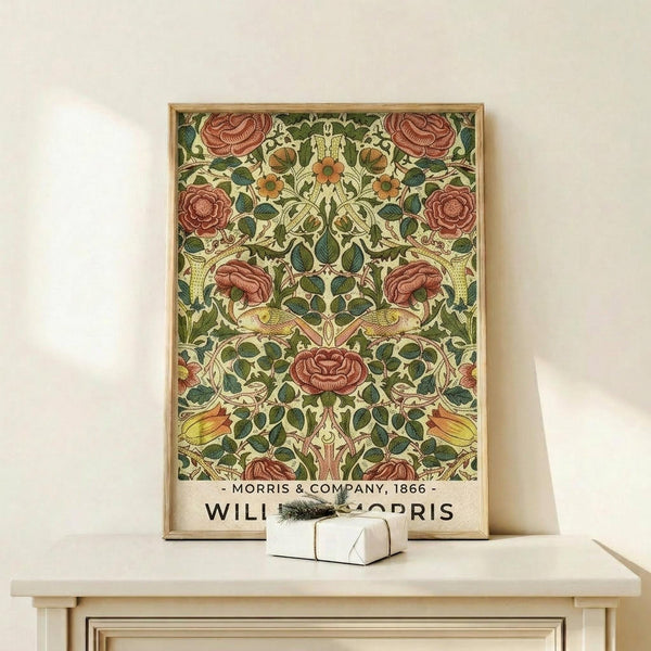 May include: Framed vintage-style botanical poster with symmetrical pattern of red roses and green foliage on cream background, displayed on light wooden console table next to small white gift box with evergreen sprig, against neutral wall in warm natural lighting.