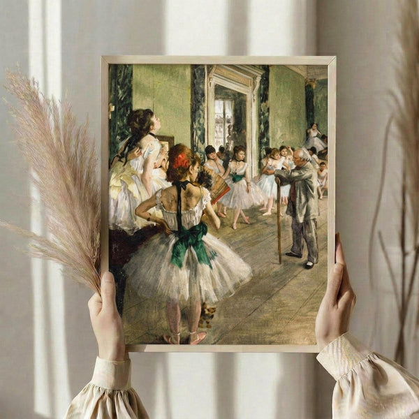 May include: Hands holding a framed reproduction of Degas' ballet class painting showing dancers in white tutus with green sashes in an ornate practice room. The print features soft greens and whites in impressionist style. Golden frame against white curtained background with dried wheat stems visible on left side.