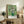 May include: Framed Van Gogh Roses poster with white flowers in green vase against pale green background, displayed on rustic wooden sideboard alongside fern plant, ceramic vases, and books in neutral-toned living room with beige chair and white walls