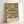 May include: Framed botanical print featuring William Morris's ornate floral pattern with coral roses, orange flowers, and green foliage in Arts and Crafts style. The artwork displays intricate symmetrical design with birds and flowing vines on cream background. Text reads "Morris & Company, 1866 - William Morris" below the pattern. Light wood frame leans against white wall with natural lighting creating soft shadows.