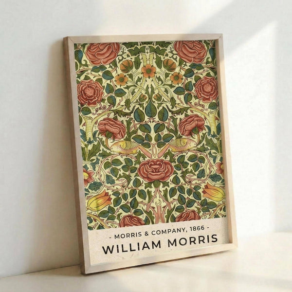 May include: Framed botanical print featuring William Morris's ornate floral pattern with coral roses, orange flowers, and green foliage in Arts and Crafts style. The artwork displays intricate symmetrical design with birds and flowing vines on cream background. Text reads "Morris & Company, 1866 - William Morris" below the pattern. Light wood frame leans against white wall with natural lighting creating soft shadows.