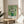 May include: Framed art print of white roses in a green vase against a light green background, displayed on a round wooden side table next to a beige upholstered chair. Natural sunlight creates shadows on the cream colored wall. The vintage-style botanical artwork features thick brushstroke textures and is matted in white with wooden frame.