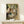 May include: Framed reproduction of Degas ballet painting showing young dancers in white tutus with green sashes practicing in an ornate room with green walls, displayed on light wood surface beside small white wrapped gift with evergreen sprig against cream wall