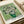 May include: Van Gogh reproduction poster print showing white roses in green vase against pale green background, displayed in wooden frame on neutral surface beside cream ribbon roll. Post-impressionist brushwork style with visible texture and swirling paint strokes throughout floral arrangement.
