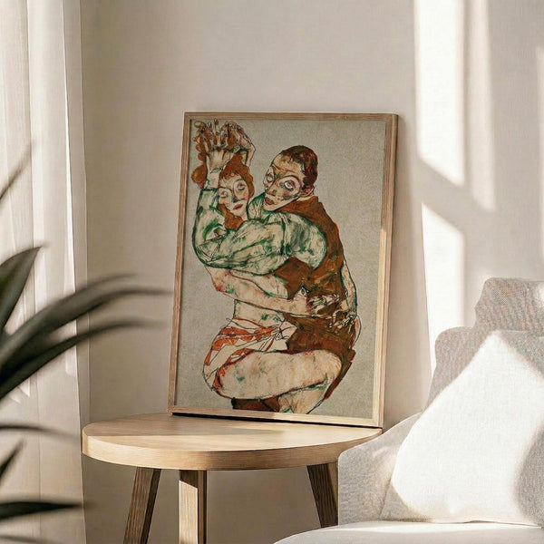 May include: Framed art print featuring two embracing figures in expressionist style with earthy tones of red, green, and brown, displayed on a round wooden side table next to a beige upholstered chair in a bright, minimalist room with white walls and natural lighting from a window.