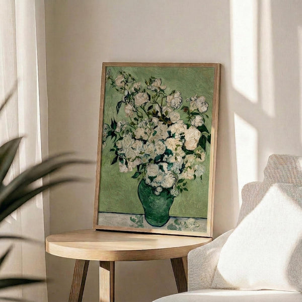 May include: Framed reproduction of Van Gogh's white roses painting displayed on round wooden side table next to beige upholstered chair. The artwork features thick white blooms in green vase against sage green background, housed in thin wooden frame. Soft natural lighting creates shadows on cream wall, with plant leaves visible in corner of modern living room setting.