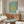 May include: Modern living room with walnut credenza displaying Matisse cut-out poster featuring yellow organic coral-like shape on turquoise background. White sculptural chair, round glass coffee table, and white table lamp complete the mid-century styled space with warm wood tones and neutral walls.