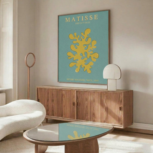 May include: Modern living room with walnut credenza displaying Matisse cut-out poster featuring yellow organic coral-like shape on turquoise background. White sculptural chair, round glass coffee table, and white table lamp complete the mid-century styled space with warm wood tones and neutral walls.