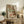 May include: Framed reproduction of an expressionist artwork showing two embracing figures in earthy tones of green, brown, and red, displayed on a rustic wooden sideboard alongside books, a ceramic vase, and a potted fern in a neutral-toned living room setting.