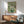May include: Van Gogh roses painting reproduction in wooden frame hanging above mid-century modern wooden credenza with white lamp, next to teal upholstered armchair with wooden arms. Artwork shows white and pink roses in green vase against sage green background. Warm sunlight creates dramatic shadows across white wall in styled living room setting.