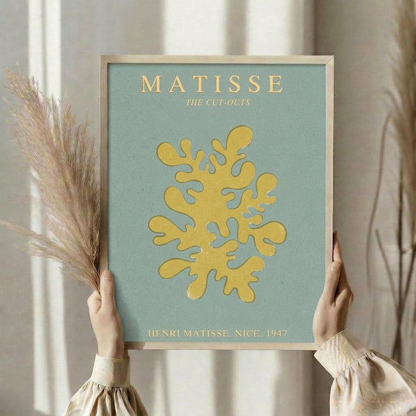 May include: Person holding framed Matisse-style poster featuring yellow organic cut-out shapes on sage green background. Golden text reads "MATISSE THE CUT-OUTS" and "HENRI MATISSE, NICE, 1947". Natural wood frame, beige curtains and dried pampas grass visible in soft-lit room setting. Person wears cream ruffle-sleeve top.