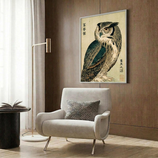 May include: Japanese woodblock print of an owl with detailed brown, teal and cream feathers displayed in silver frame on wood-paneled wall above beige upholstered chair with geometric patterned pillow, next to brass floor lamp and dark round side table with open book
