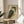 May include: Framed Japanese woodblock print of an owl with detailed brown and teal feathers, bright orange eyes, displayed on a round wooden side table next to a beige upholstered chair. Natural sunlight streams across the neutral-toned room setting, creating soft shadows on white walls.