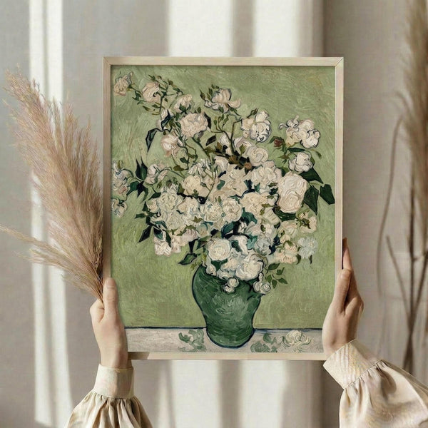 May include: Hands holding a framed reproduction of Van Gogh's painting showing white roses in a green vase against a pale green background, displayed in a light wooden frame. Person wearing cream-colored clothing with gathered sleeves holds dried pampas grass in one hand while positioning the artwork near white curtains in soft natural lighting.