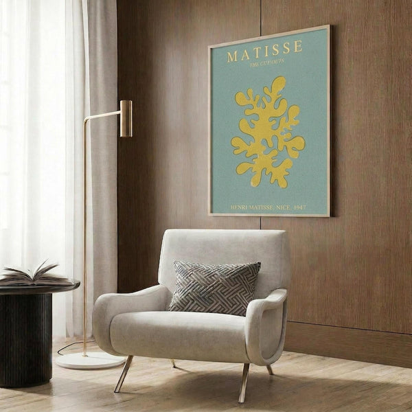May include: Matisse poster print with yellow coral-like cutout design on sage green background, displayed in modern reading nook with beige upholstered chair, geometric throw pillow, brass floor lamp, and wood-paneled wall. Frame appears light wood with clean lines.