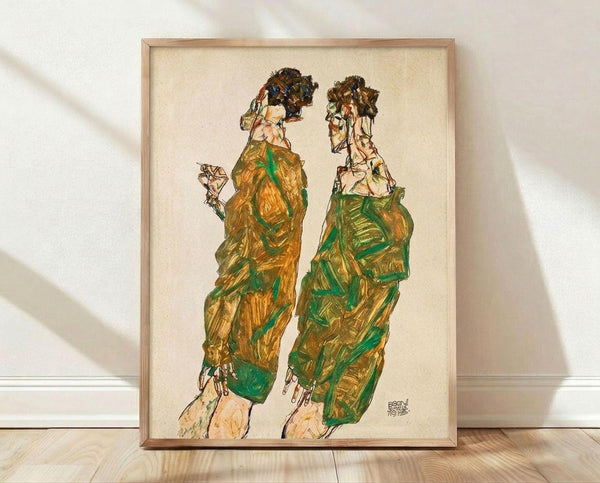 May include: Framed reproduction of an expressionist drawing showing two elongated figures in flowing green and golden robes with dark hair, rendered in sketchy lines against cream paper. The wooden frame sits on light hardwood flooring against a white textured wall with diagonal shadows.