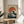 May include: Framed poster print of Hokusai's iconic Great Wave with large red sun, displayed on small round wooden side table next to beige upholstered chair. Natural sunlight streams across white walls creating geometric shadows. Green plant leaves visible in corner. Print shows dramatic blue and white ocean waves against cream background in thin light wood frame.