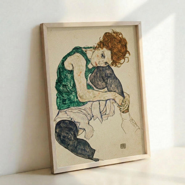 Framed art print showing a seated woman with curly reddish hair wearing a green sleeveless top and dark stockings. The expressionist-style drawing features bold outlines and muted colors on cream paper. The wooden frame leans against a white wall, casting soft shadows. The artwork displays characteristic early 20th century Austrian art style with angular forms and emotional intensity.