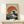 May include: Framed Japanese-inspired art print featuring large ocean wave with white foam crashing beneath orange-red circular sun on cream speckled background, displayed on light wood surface with small white wrapped gift box, leaning against white wall with natural lighting and shadows