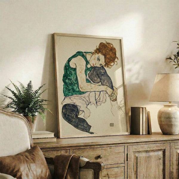 May include: Framed art print featuring a seated woman with reddish curly hair wearing a green sleeveless top and dark stockings, displayed on a rustic wooden sideboard alongside books, a small potted fern, and ceramic vase in a neutral-toned living room setting with cream upholstered furniture