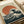 Japanese woodblock print style artwork featuring the iconic great wave with dramatic curling blue-green waves and white foam against a large orange-red circular sun in the background. The print has a cream colored background with traditional Japanese text in the upper corner. The artwork is displayed on white paper with rolled paper visible in the background, showing the vintage ukiyo-e art style with detailed line work and classic color palette.