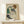 May include: Framed reproduction of an expressionist drawing showing a seated woman with curly auburn hair wearing a green sleeveless top and dark stockings, rendered in sketchy lines with muted colors on cream paper. The wooden-framed artwork leans against a white wall on a light wood surface next to a small white wrapped gift with evergreen sprig.