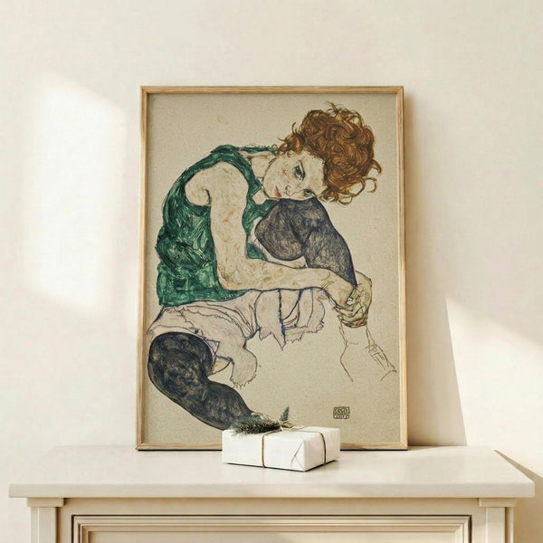 May include: Framed reproduction of an expressionist drawing showing a seated woman with curly auburn hair wearing a green sleeveless top and dark stockings, rendered in sketchy lines with muted colors on cream paper. The wooden-framed artwork leans against a white wall on a light wood surface next to a small white wrapped gift with evergreen sprig.
