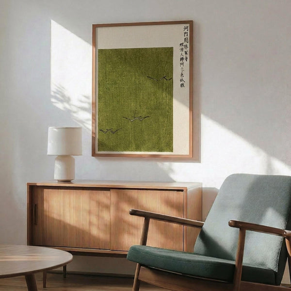 May include: Framed Japanese artwork with green textured background and three flying birds in black ink, displayed above a wooden sideboard with table lamp in modern living room setting with sage green upholstered chair and white walls with natural sunlight shadows.