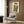 May include: Framed art print displaying two elongated figures in green and brown garments against cream background, hanging on wood-paneled wall above beige upholstered chair with geometric patterned pillow, brass floor lamp and dark round side table with open book nearby in modern interior setting.