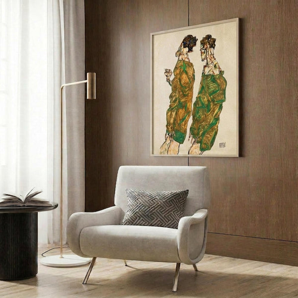 May include: Framed art print displaying two elongated figures in green and brown garments against cream background, hanging on wood-paneled wall above beige upholstered chair with geometric patterned pillow, brass floor lamp and dark round side table with open book nearby in modern interior setting.