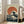 May include: Framed Japanese-style artwork featuring a large dark blue ocean wave with white foam against a bright orange-red circular sun on a cream background with speckled texture, displayed in a light wood frame leaning against white wall next to beige upholstered chair in bright living room with hardwood floors and large windows