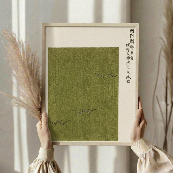 May include: Person in cream ruffled blouse holding framed artwork against white curtained window, with dried wheat stems. Green textured poster features minimalist black line drawings of flying birds and vertical Japanese calligraphy text on cream background in light wood frame.