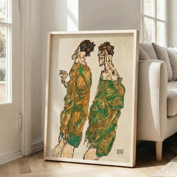 May include: Large framed art print leaning against white wall in bright living room with beige sofa. Artwork shows two figures in flowing green and brown robes with dark hair, rendered in expressive sketchy style with visible brushstrokes. Light wood frame surrounds cream-colored print. Natural light streams through tall windows onto hardwood floors.