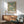 May include: Framed botanical poster with pink chrysanthemums and green foliage pattern hanging above wooden sideboard in modern living room. Mid-century sage green armchair with wooden arms sits beside credenza topped with ceramic table lamp. Natural sunlight creates shadows on white wall, showing vintage floral wallpaper design in earth tones with blue accents.