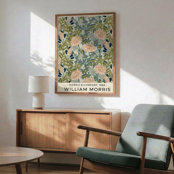 May include: Framed botanical poster with pink chrysanthemums and green foliage pattern hanging above wooden sideboard in modern living room. Mid-century sage green armchair with wooden arms sits beside credenza topped with ceramic table lamp. Natural sunlight creates shadows on white wall, showing vintage floral wallpaper design in earth tones with blue accents.