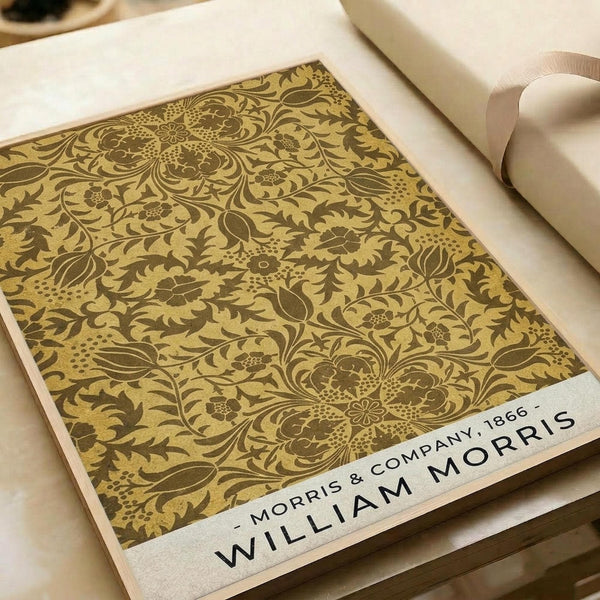 Ornate floral wallpaper pattern in golden yellow and brown tones featuring intricate Victorian-style botanical motifs with swirling vines, detailed flowers, and decorative leaves. The design sample is displayed on white paper with Morris & Company 1866 attribution text at bottom, photographed alongside rolled white papers on a neutral surface.