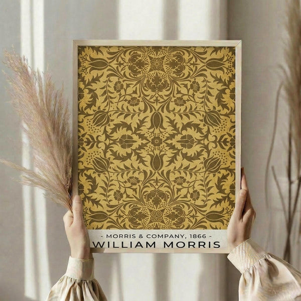 May include: Person in beige sweater holding framed poster featuring ornate golden botanical damask pattern with intricate leaves, flowers and symmetrical design. Dried pampas grass visible beside figure against soft neutral curtained background in natural lighting.