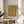 May include: Framed vintage William Morris floral pattern print in golden yellow and brown tones displayed on a round wooden side table next to a beige upholstered chair. Natural sunlight streams through white curtains creating soft shadows on the neutral wall. A green plant leaf is partially visible in the corner, adding warmth to the minimalist room setting.