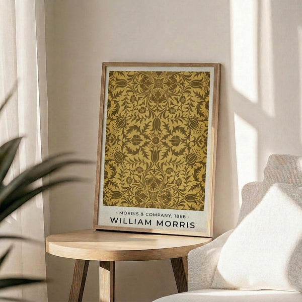 May include: Framed vintage William Morris floral pattern print in golden yellow and brown tones displayed on a round wooden side table next to a beige upholstered chair. Natural sunlight streams through white curtains creating soft shadows on the neutral wall. A green plant leaf is partially visible in the corner, adding warmth to the minimalist room setting.