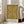 May include: Framed art print leaning against white wall next to beige upholstered chair in bright living room with hardwood floors and large windows. The poster features an ornate golden yellow and brown Victorian floral damask pattern with intricate botanical motifs, leaves, and symmetrical designs. White text at bottom reads "Morris & Company, 1866 - William Morris" on clean white background within light wood frame.