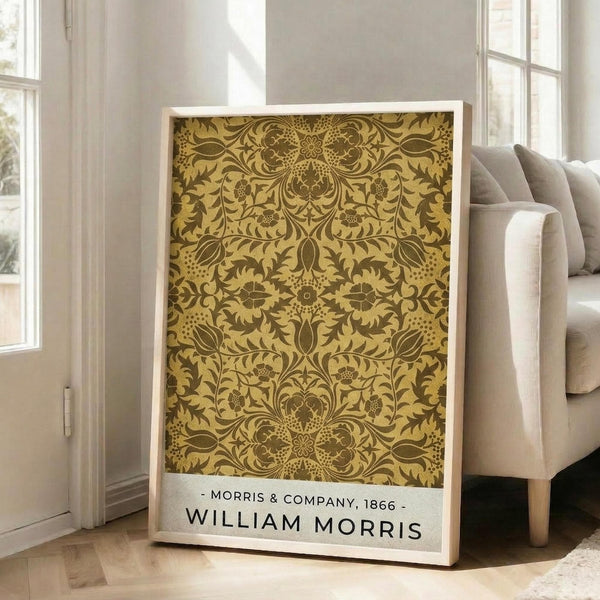May include: Framed art print leaning against white wall next to beige upholstered chair in bright living room with hardwood floors and large windows. The poster features an ornate golden yellow and brown Victorian floral damask pattern with intricate botanical motifs, leaves, and symmetrical designs. White text at bottom reads "Morris & Company, 1866 - William Morris" on clean white background within light wood frame.