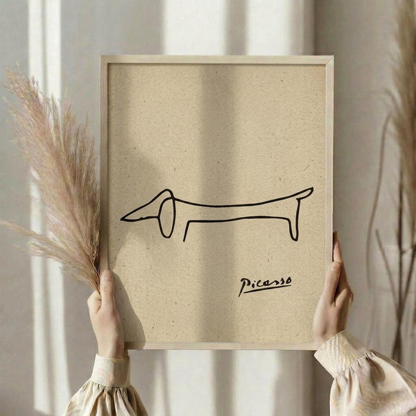 May include: Hands holding a framed minimalist line drawing of a dachshund dog on beige textured paper, signed "Picasso" in black ink. The simple continuous line art shows the dog's elongated body and droopy ears in profile. Natural wood frame, neutral beige clothing, dried pampas grass visible on left side, soft window lighting in background.